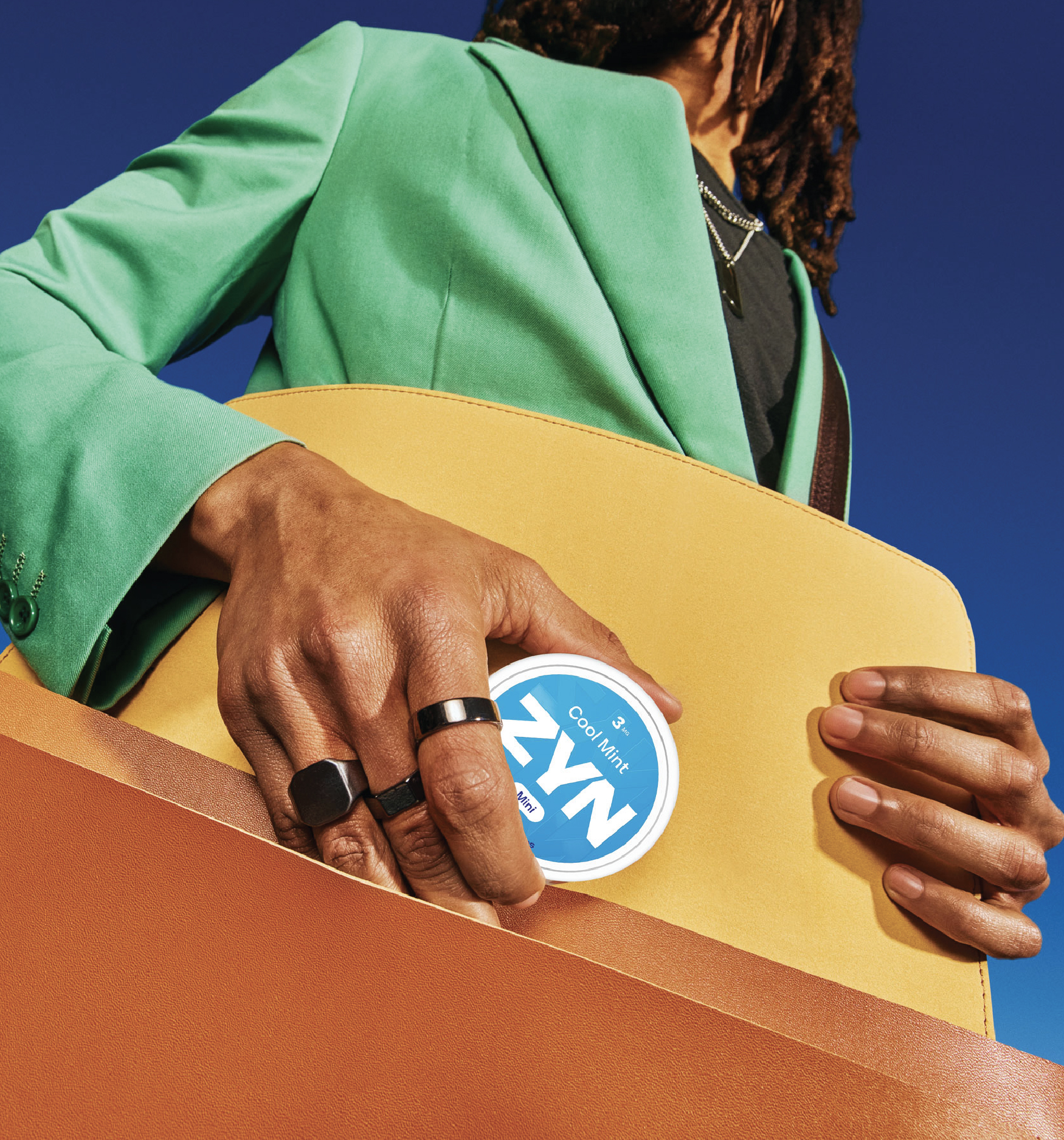 A person in a green suit placing a white ZYN nicotine pouches container inside an orange satchel behind a blue background.